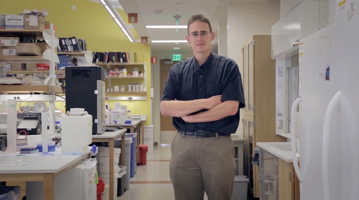 Rob Knight is pictured, arms-folded, standing in a laboratory surrounded by equipment and apparatus.