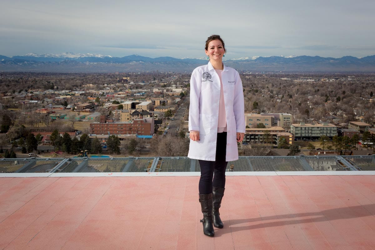 Roberta Capp is pictured standing on a rooftop with a picturesque view of the city and the Rocky Mountains behind her.