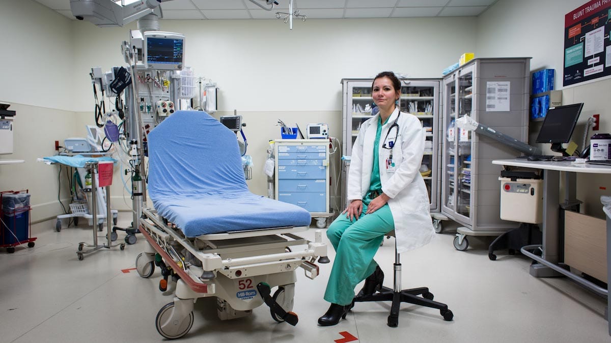 Roberta Capp is pictured seated alongside a portable hospital bed fitted with equipment, wearing scrubs and a white lab coat.