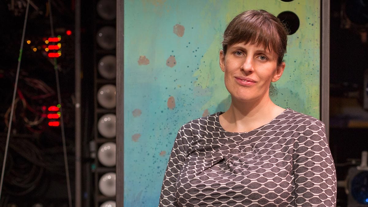 Sarah Benson is pictured from the chest-up, wearing a printed shirt, standing in front of a green and blue painted stage piece.