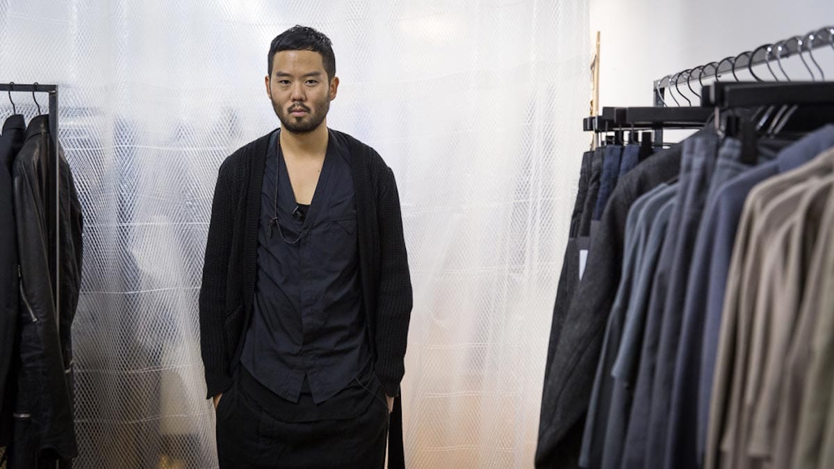 Siki Im, dressed in a long black sweater, stands alongside garments hanging on clothing racks.