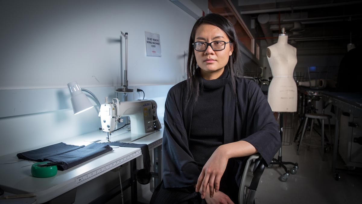 Tuyen Tran sits in front of a sewing machine, wearing a black sweater and glasses, with a mannequin in the background.