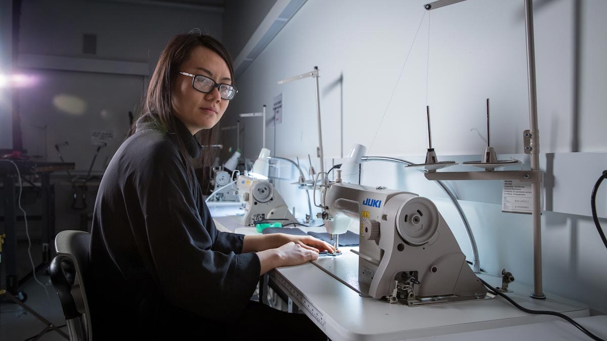 Tuyen Tran, wearing a black sweater and glasses, sits sewing using a sewing machine.