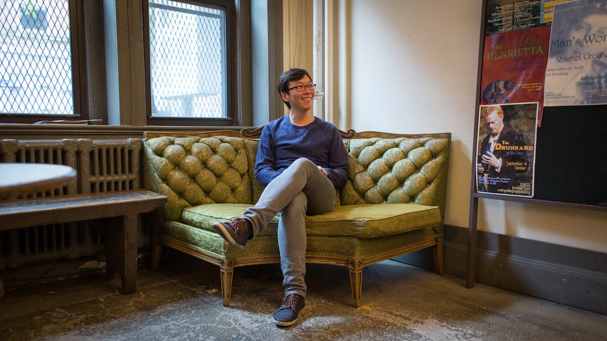 Yi Zhao is pictured smiling and sitting with his legs crossed on a green victorian-style sofa with theater advertisements tacked to a board alongside him.