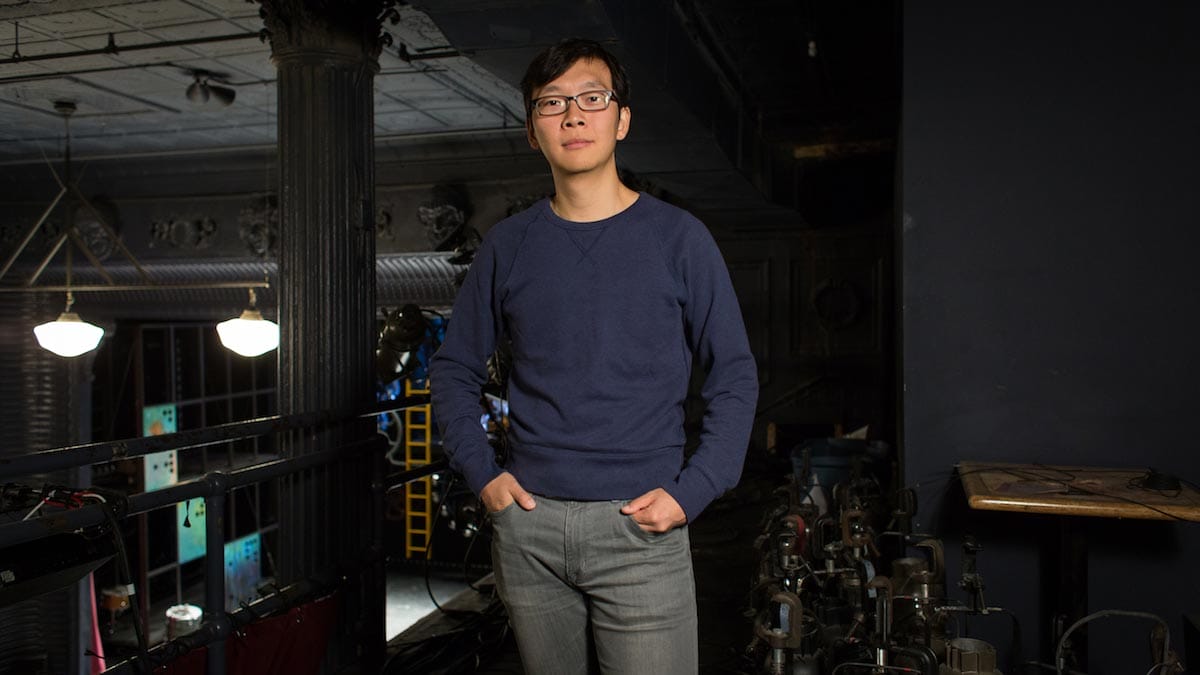 Yi Zhao is pictured standing on a balcony of a theater, with lighting equipment behind him and production equipment visible on the stage.