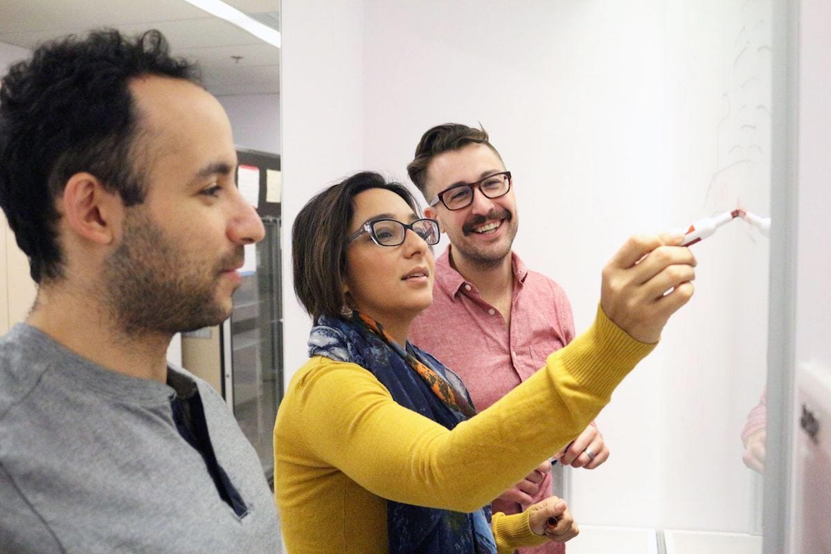 Houra Merrikh is pictured wearing a yellow sweater, standing between two students looking at a white board as she writes on it with a marker.