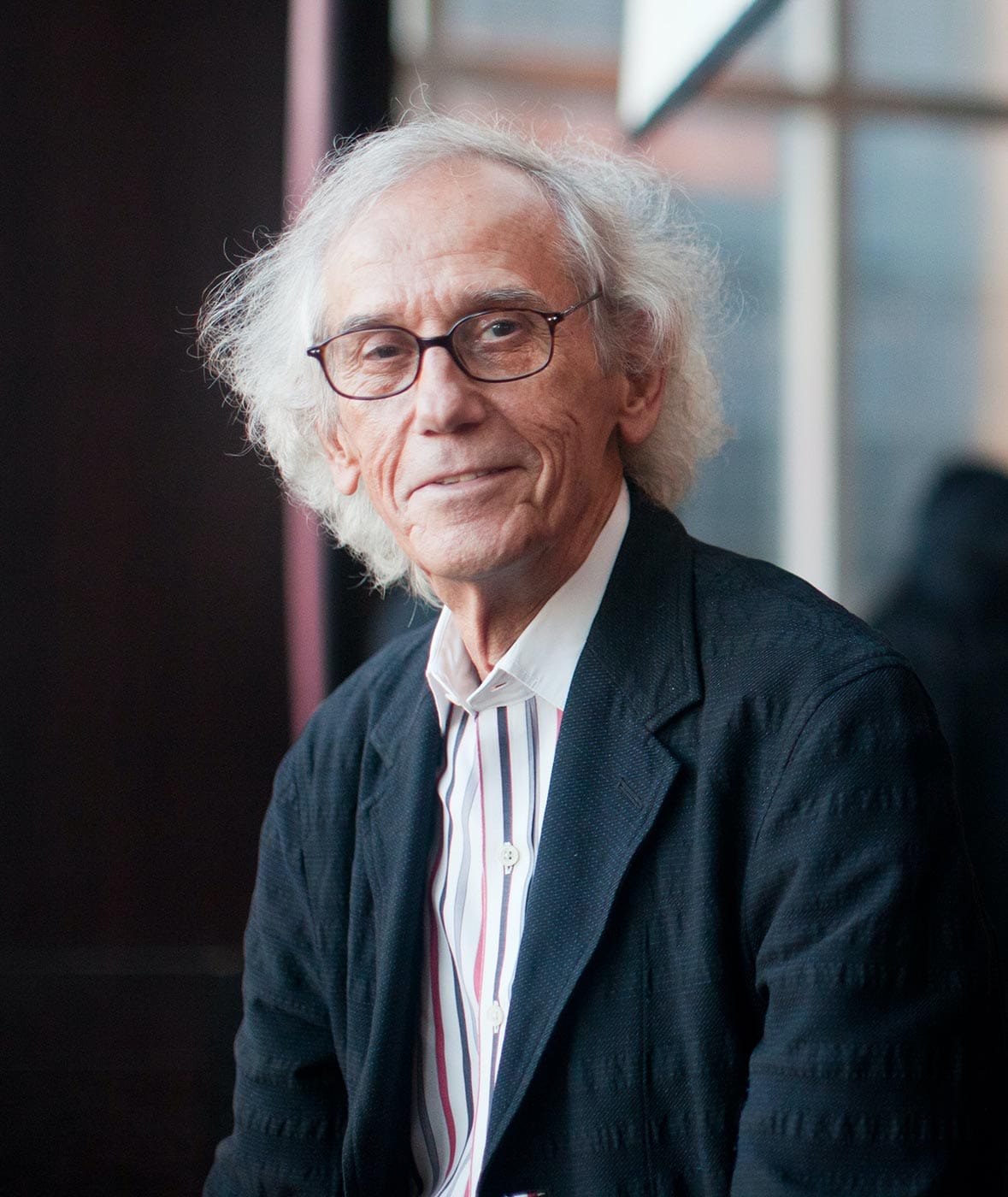 Christo standing in front of a wall of windows at the Mandarin Oriental in New York City.