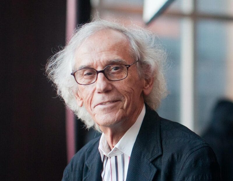 A photo of Christo in front of a wall of windows.