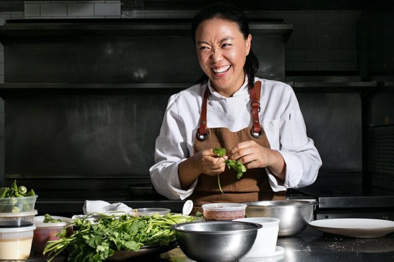 Chef Jae is pictured standing in a kitchen with various ingredients in front of her as she wears an apron and an animated smile on her face.