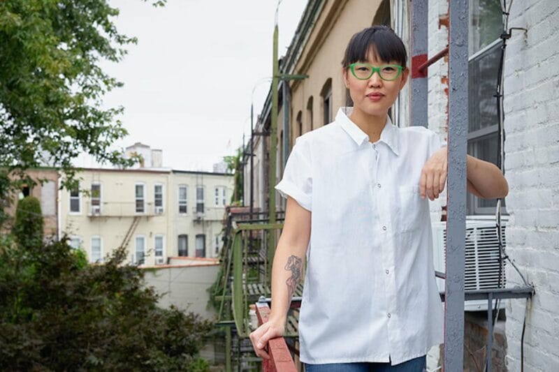 Chef Kate is pictured standing on a fire escape, wearing a white blouse and green thick-rimmed glasses.