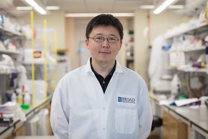 Feng Zhang is standing in an aisle in his lab, dressed in a white lab coat.