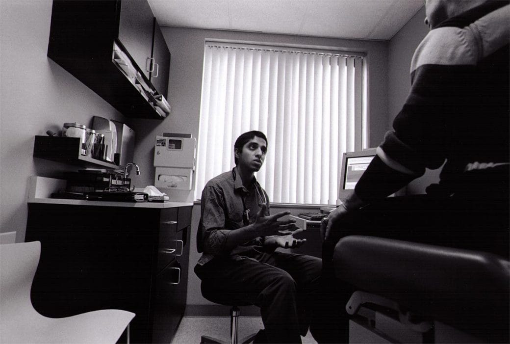 Black and white photograph of Vivek Murthy, sitting in a doctor's office, speaking to a patient.