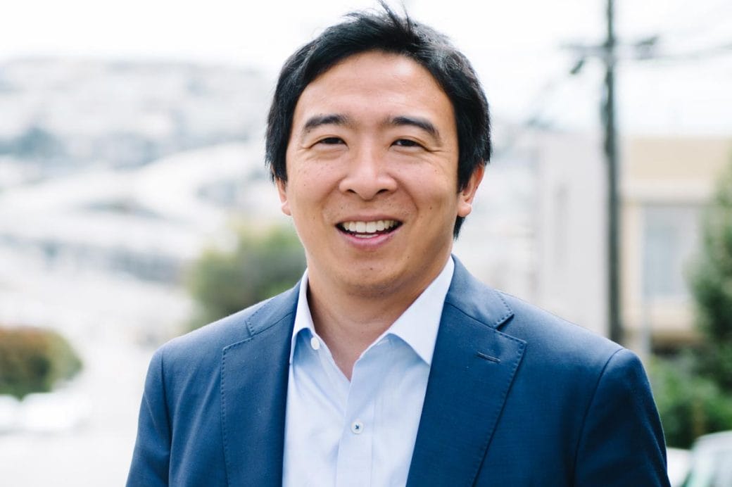 Andrew Yang, in a blue suit jacket and shirt, standing outside with a city in the background.