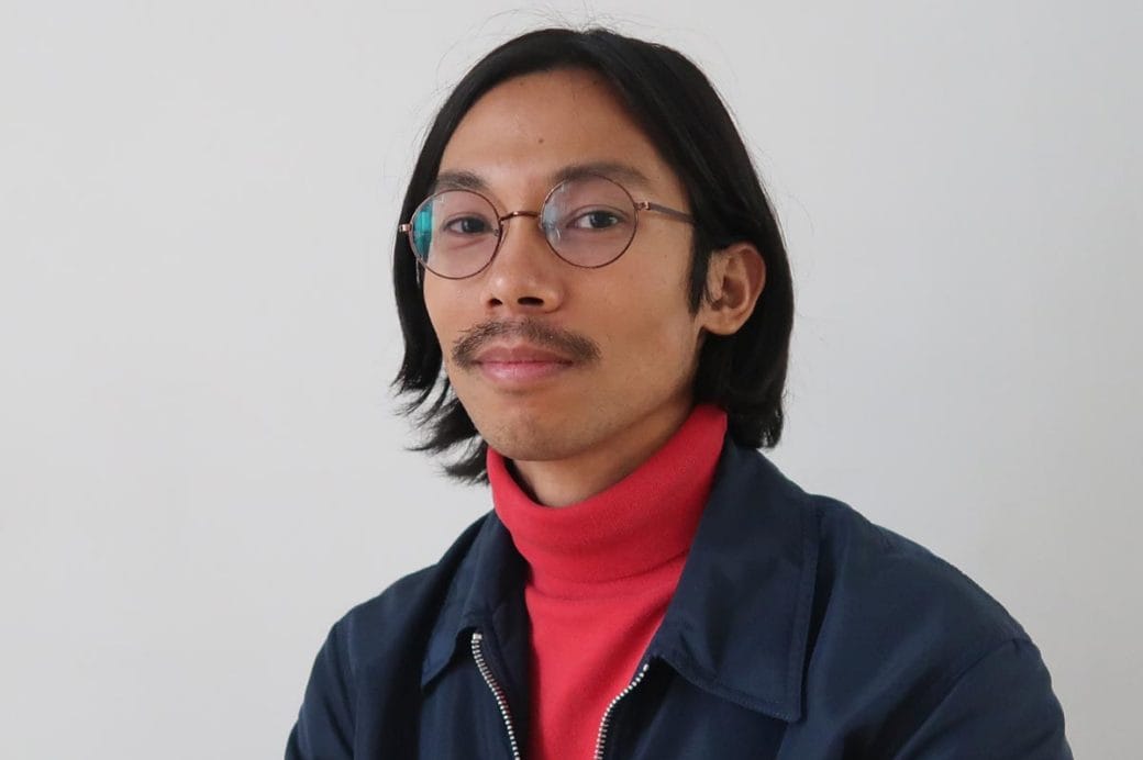 Miko Revereza, wearing a blue jacket and red turtleneck, sitting against a neutral gray background.