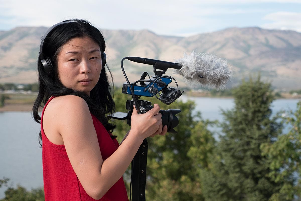 Nanfu Wang, holding a camera and microphone, filming on location in Florida with trees, mountains, and water in the distance.