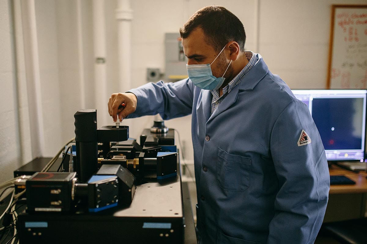 Mohamed, wearing a lab coat and mask, adjusts lab equipment.