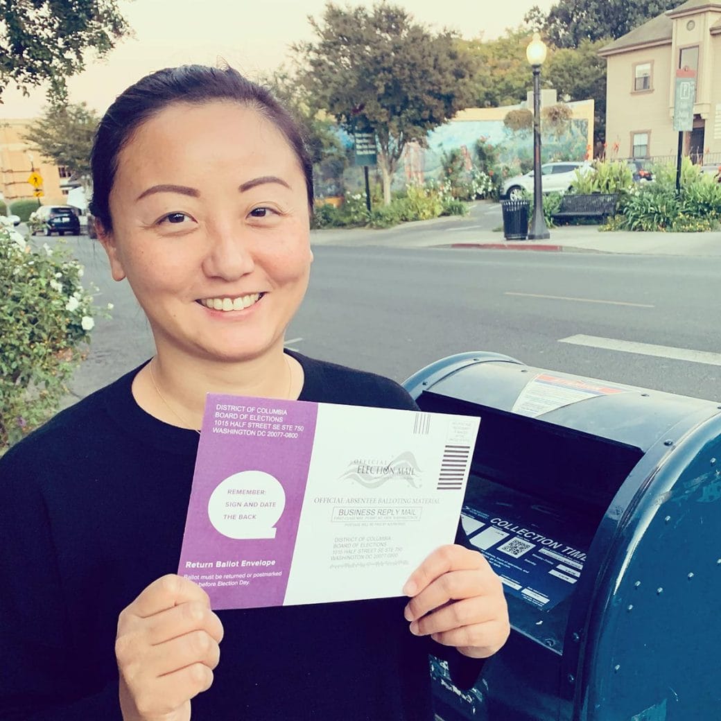 Yi Chen holding up a mail-in ballot in front of a USPS mailbox.