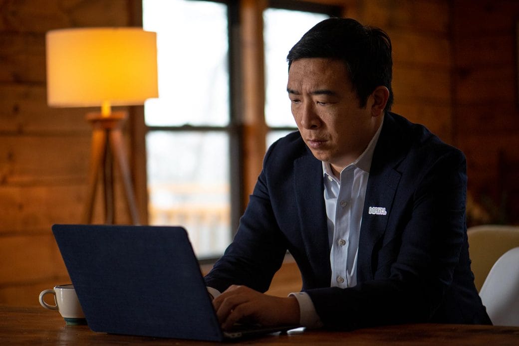 Andrew Yang, in blue suit jacket and MATH pin, sitting at a table typing on a laptop.