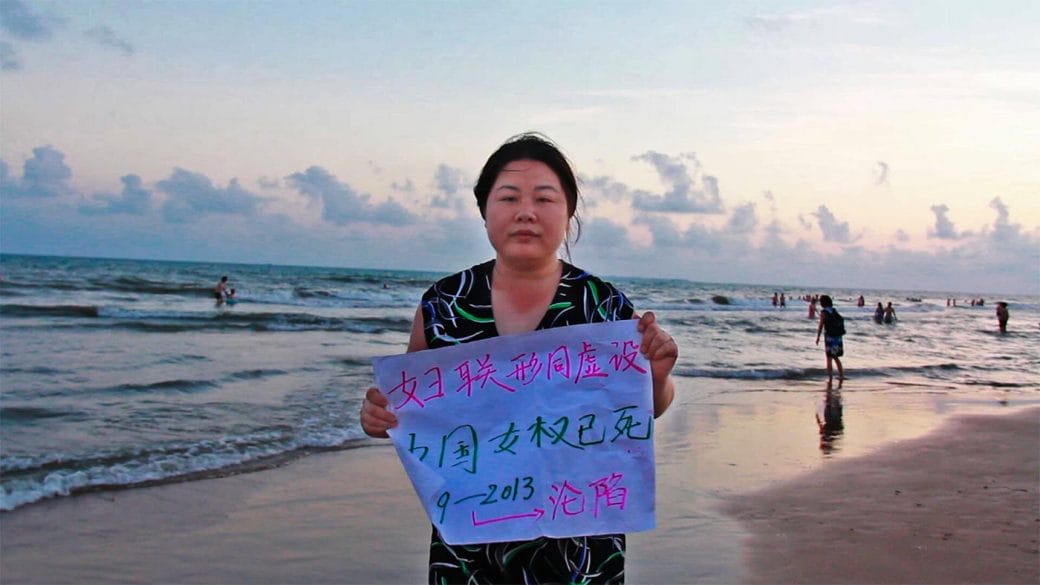 A still frame of Chinese activist Ye Haiyan holding a sign on a beach, from the film Hooligan Sparrow.