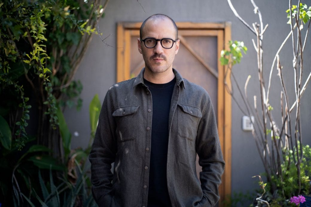 Juan Pablo wearing thick-rimmed glasses and a button down shirt framed by fauna.