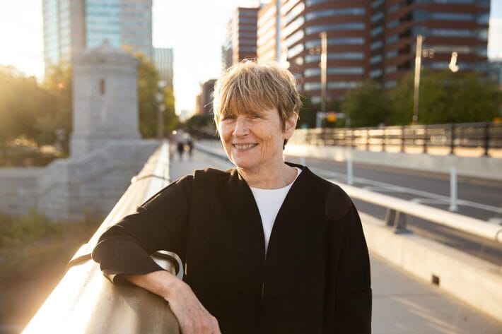 A portrait of Ruth Lehmann in a black blazer as she stands on a bridge over the Charles River.