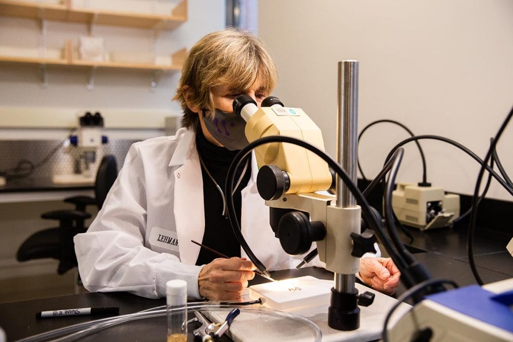 Ruth Lehmann in a white lab coat, sitting in front of a microscope analyzing a sample.