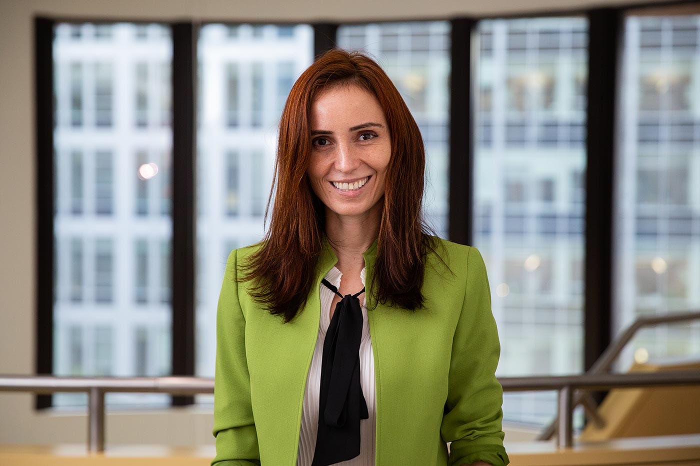 Silvi Rouskin, in a green blazer, standing in front of a wall of windows and stairwell in MIT.