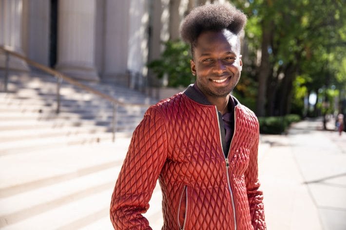 Ibrahim Cissé with a big smile, wearing a red jacket, standing in front of the steps of an MIT building.