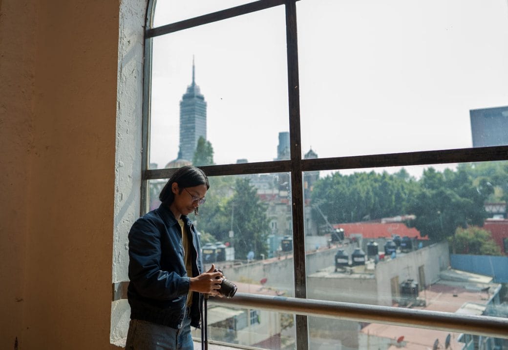 Miko Revereza stands in front of a large window with a view of the city behind him.