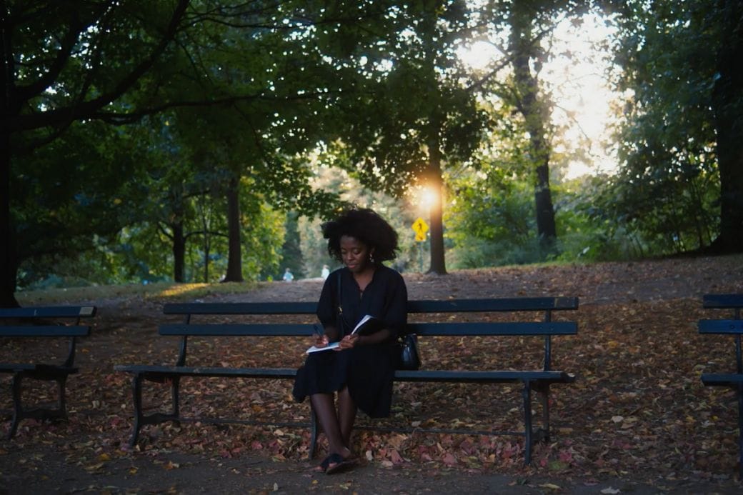 Yaa Gyasi sits on a park bench reading surrounded by green trees. 