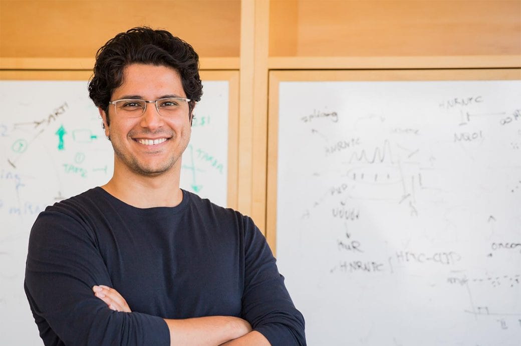 Hani Goodarzi, arms crossed, standing in front of a whiteboard with scientific equations.