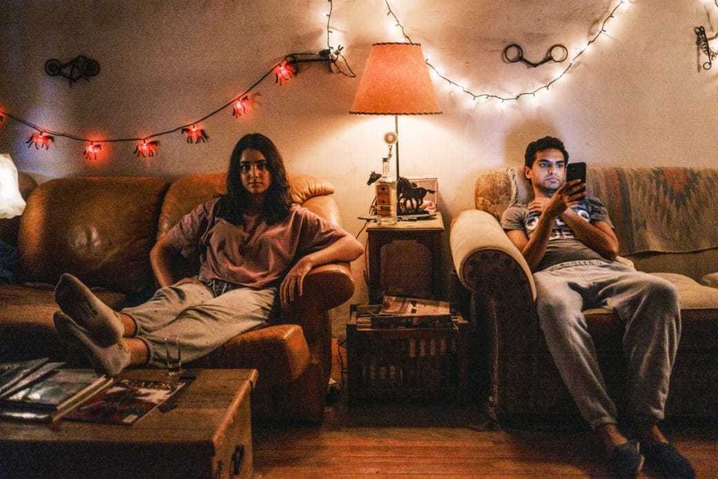 Rita and Ravi sit on separate chairs within the living room of an apartment.