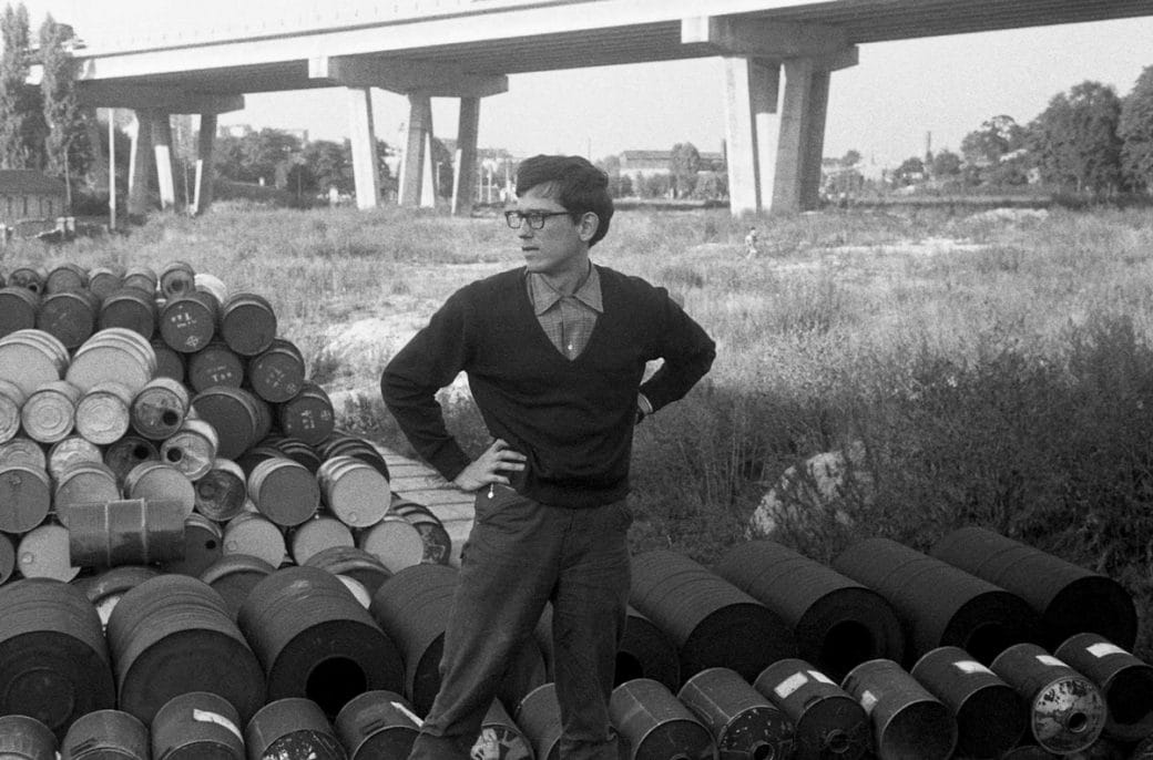 A young Christo in the 1950s standing in front of a mound of oil barrels.