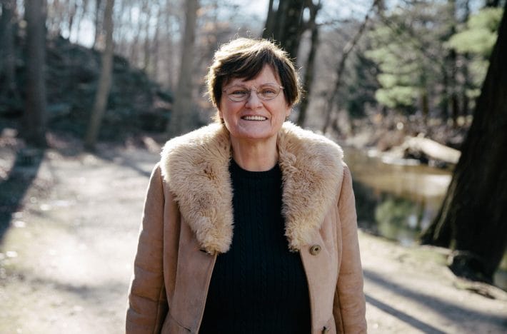 Katalin Karikó standing in the woods in a beige coat with fur trim.