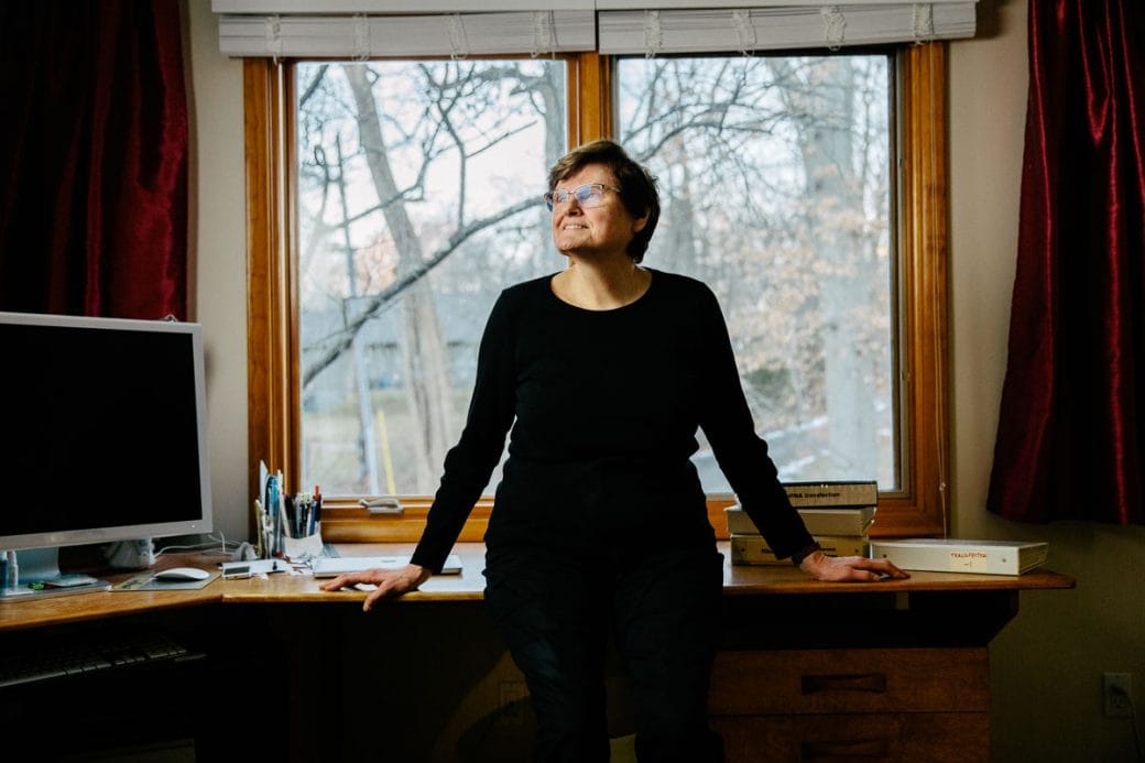 Katalin Karikó in all black leaning on her desk and looking up to the left.