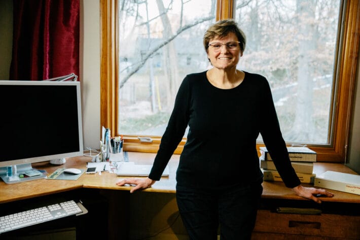 Katalin Karikó in all black standing with her hands on her desk, smiling.