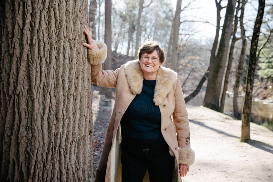 Katalin Karikó in a fur-collared jacket smiling and leaning against a tree trunk.