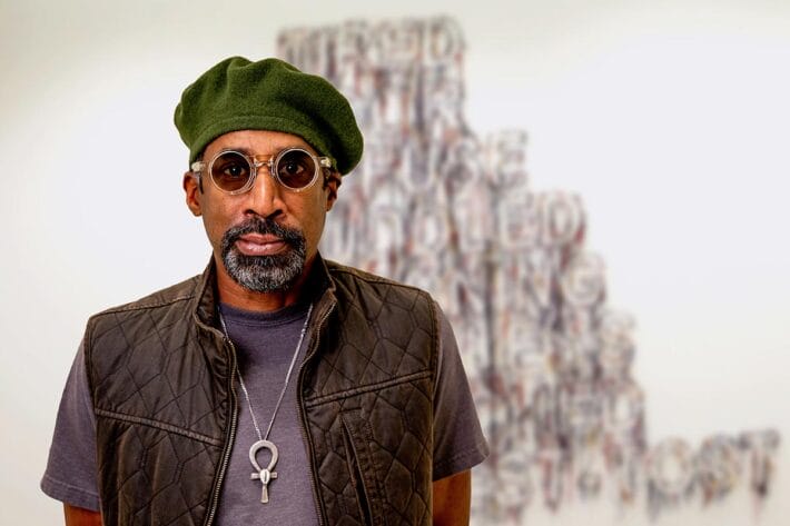 Nari Ward, wearing sunglasses and vest, stands in front of an art installation.