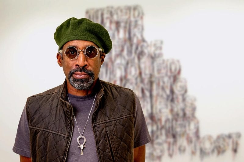Nari Ward, wearing sunglasses and vest, stands in front of an art installation.