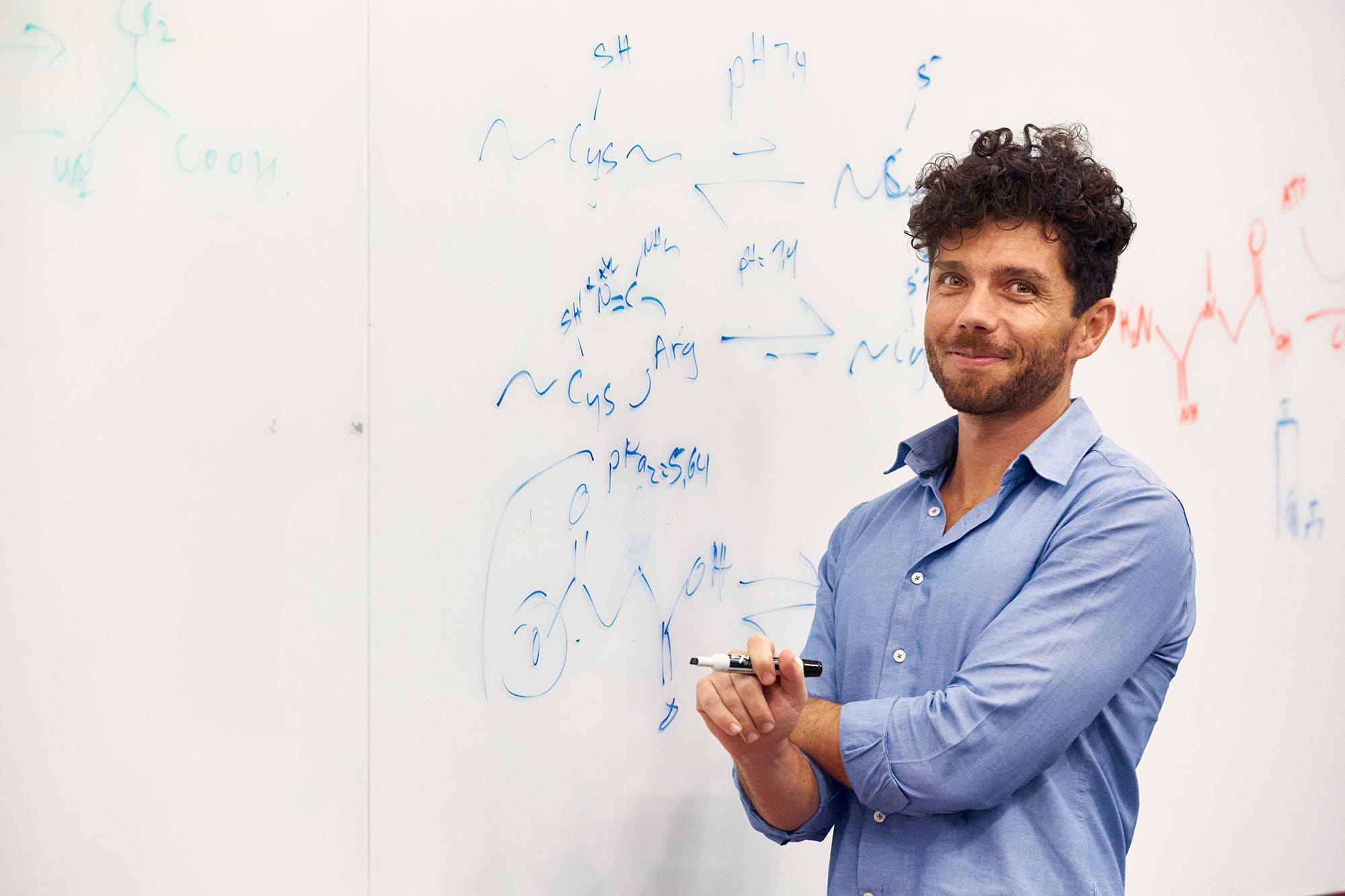 Edward Chouchani in a blue dress shirt stands in front of a whiteboard.