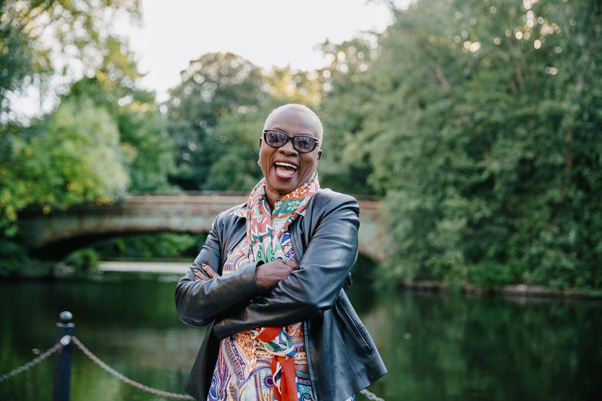 Angélique Kidjo posing for the camera and laughing in front of a small lake and a bridge.