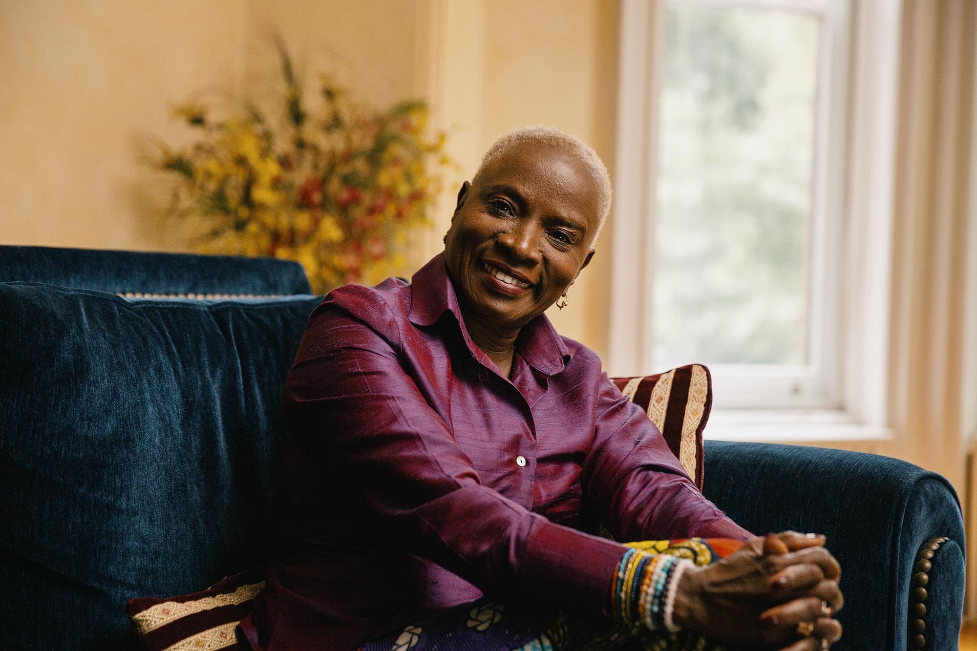 Angélique Kidjo, hands on knees, leaning forward and smiling while sitting on a couch.