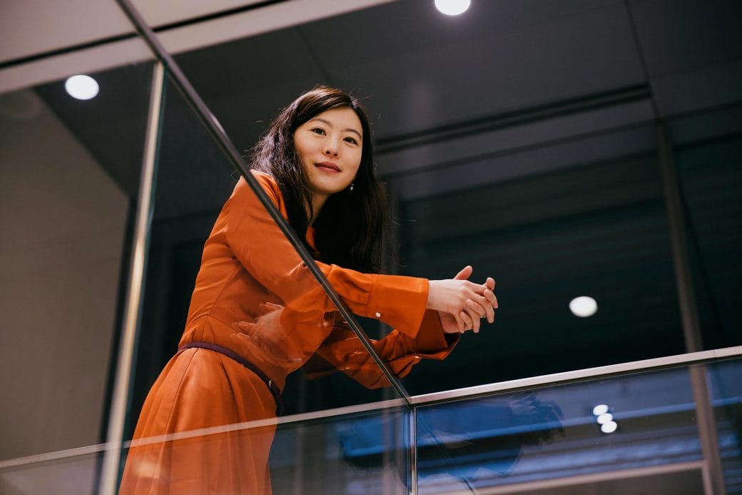 Biyu J. He wearing an orange dress and looking over a railing.