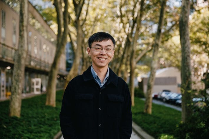 Shixin Liu wearing a black sweater framed by the tree trunks behind him.