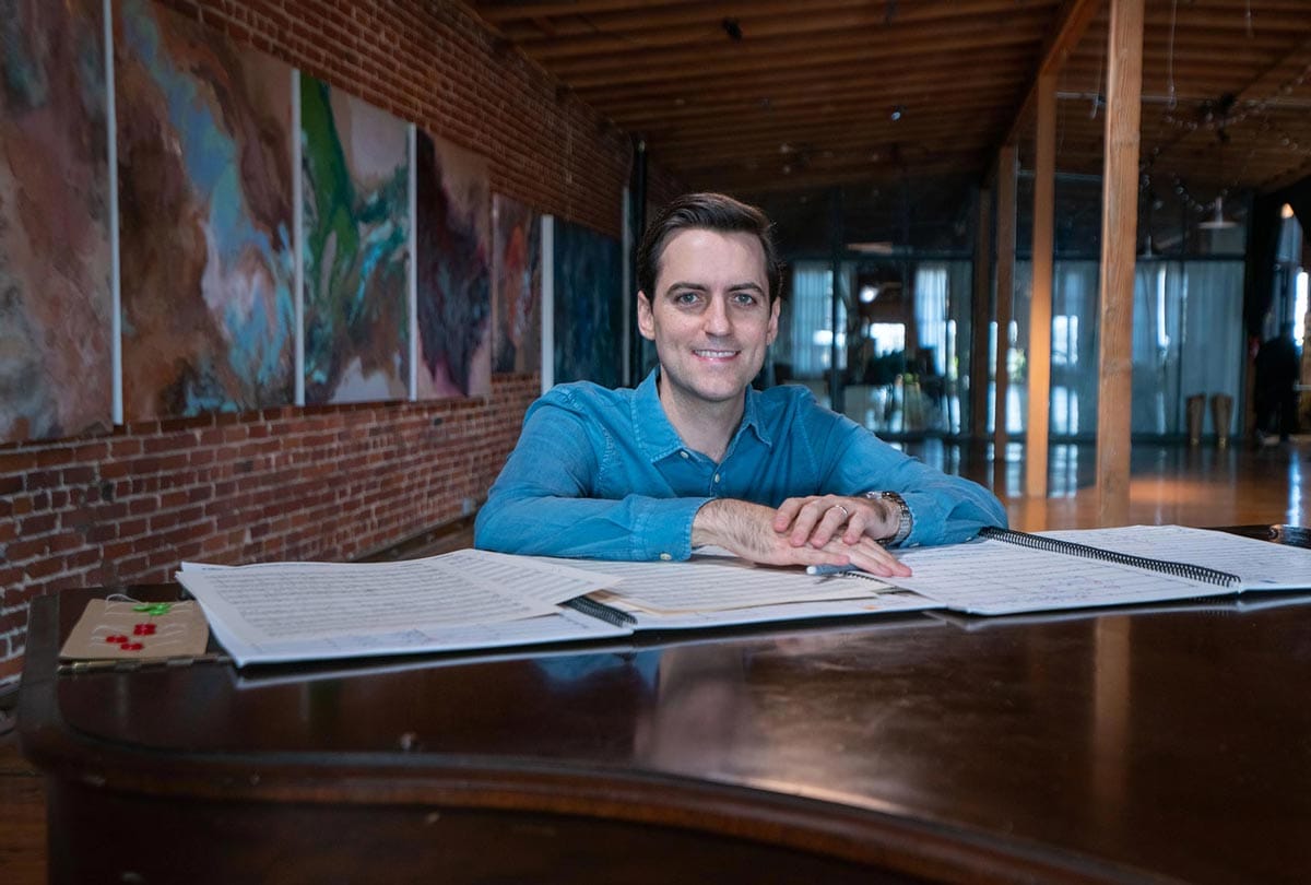 Juan Pablo Contreras stands at a piano with sheet music spread out in front of him and abstract paintings behind him.