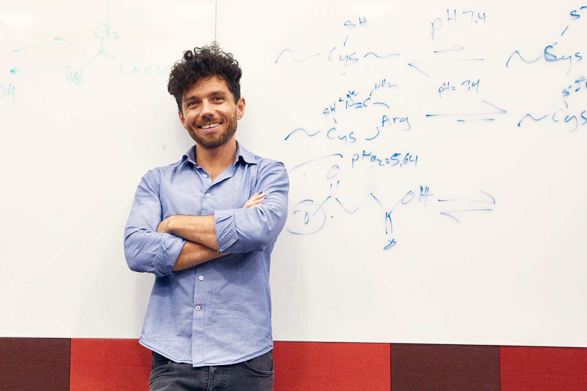 Edward Chouchani with his arms folded in front of a white board with chemistry notes on it.