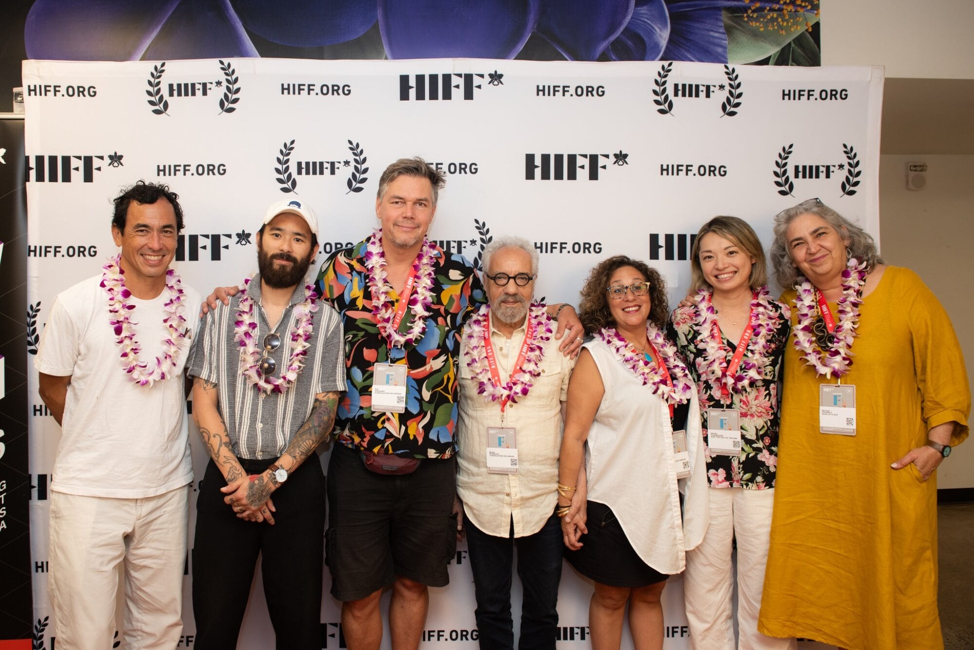 A group of seven adults, wearing leis, stand in front of a white step-and-repeat banner with HIFF and HIFF.ORG logos. From left to right: a man with dark, curly hair and a white shirt; a man with a beard and a striped shirt; a man with short, graying hair and a colorful patterned shirt; a man with a beard and glasses wearing a light-colored shirt; a woman with curly hair and a white top; a woman with dark hair and a floral patterned top; and a woman with graying hair and a yellow dress. They are all smiling.