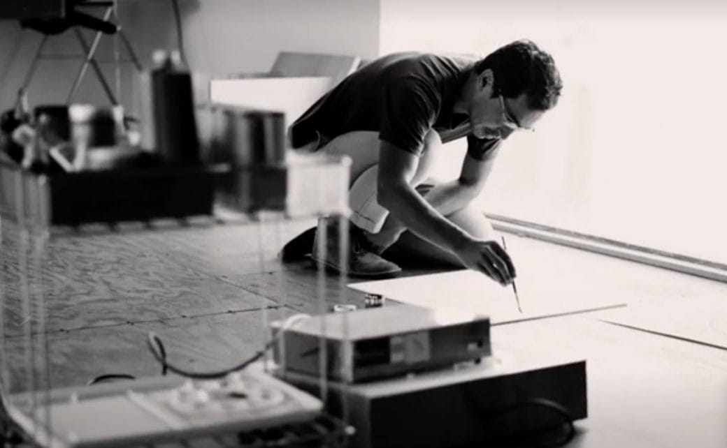 A black and white photograph of artist Geoff McFetridge crouching over a painting.