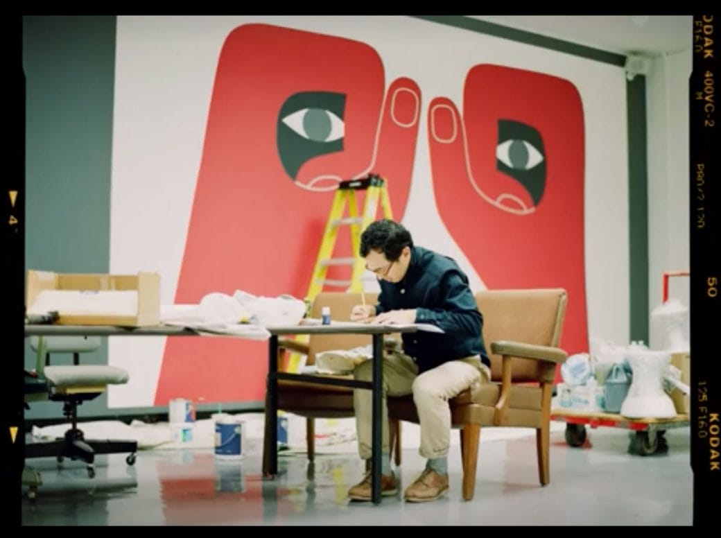Artist Geoff McFetridge working in a gallery space with a mural behind him.