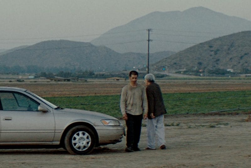 A still with two men standing at the front of an old gold car.